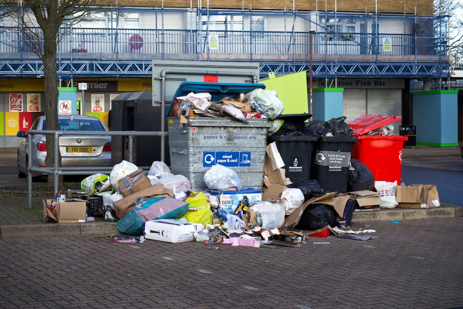 A large collection of bulky household waste items, including cardboard boxes, black plastic bin bags, and loose packaging materials, accumulated around a collection bin in an urban setting. The waste appears to comprise discarded furniture, appliances, and packaging, with some items partially on the pavement and others leaning on the metal railing nearby. Behind the waste, there is a parked silver car and a commercial building with a yellow storefront, signage, and a second-floor metal balcony with blue safety netting. The scene is set during daytime with natural light, indicating a typical urban environment where waste disposal and home relocation services by Man With a Van Rotherhithe may be involved for proper furniture and appliance disposal, in line with the page title regarding bulky waste and moving logistics.