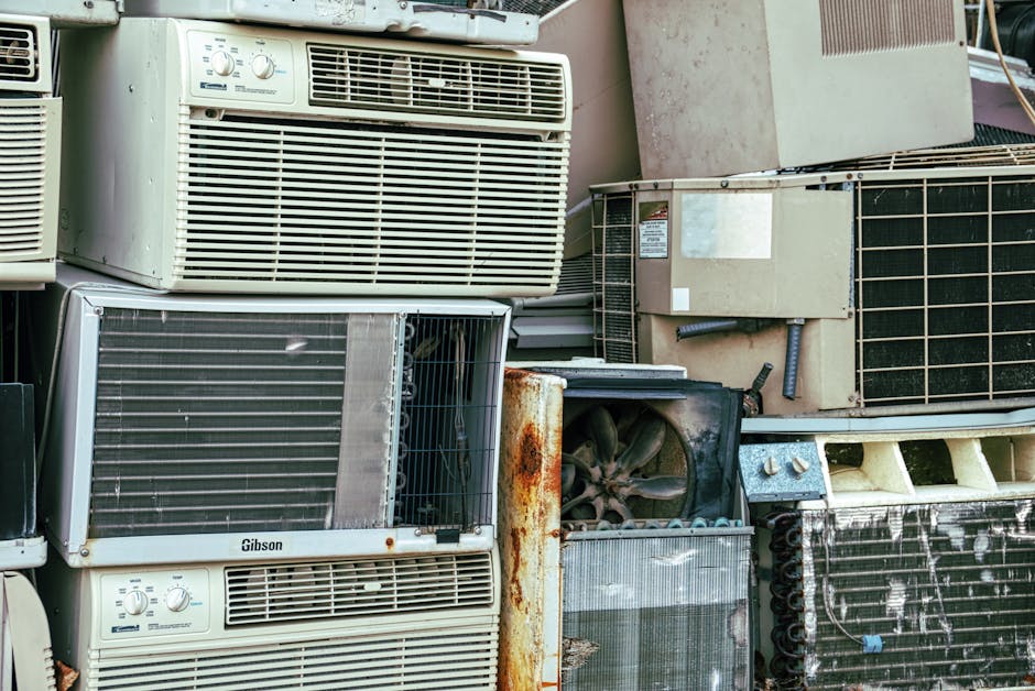 A collection of old and discarded household appliances and furniture stacked outdoors, including air conditioning units, radiators, and rusty metal parts. Some items show signs of corrosion and wear, with visible rust and dirt. The appliances are arranged on a paved surface next to a building, with some positioned inside an open van or loading area as part of a home relocation or furniture transport process. The image emphasizes the need for bulky waste disposal services, such as those offered by Man With a Van Rotherhithe, which specializes in removals and clearance of large household items in the SE16 area.