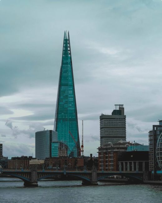 A tall, modern glass skyscraper with a pointed top dominates the skyline, situated behind a bridge spanning a river in an urban area. The sky above is overcast with grey clouds, and reflections of nearby buildings are visible in the river below. In the foreground, the bridge features multiple arches and a roadway, with no visible vehicles or pedestrians. To the right, portions of other buildings, including a high-rise structure with a flat roof and a smaller building with a rounded window feature, are visible. The scene captures the typical cityscape setting associated with professional home relocation and moving logistics in London’s Southwark area, emphasizing the urban environment where furniture transport and packing may occur.