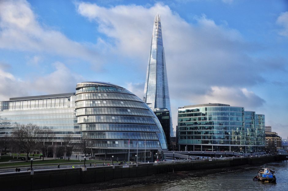 A cityscape view featuring the modern London City Hall building with its rounded, glass facade and a sleek, curved design, located alongside the River Thames. Behind City Hall, the tall, pointed Shard skyscraper rises into the partly cloudy blue sky, with reflective glass windows catching the sunlight. In the foreground, the river is visible with a small boat navigating the water. The image captures a portion of the riverside area, with pedestrians walking along the pavement and a few trees lining the walkway. The scene is well-lit with natural daylight, showcasing a typical urban environment suitable for house removals, furniture transport, and home relocation logistics, with buildings indicating a central London location. Man With a Van Rotherhithe occasionally undertakes packing and moving services in this area, supporting efficient relocation processes.