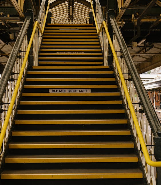 A close-up view of a stairway with black and yellow striped steps, situated in a multi-level industrial or commercial building. The staircase has yellow handrails on both sides, and a sign on the middle step reads 'PLEASE KEEP LEFT.' The surrounding environment features visible metal beams, pipes, and structural elements indicative of a warehouse or building under renovation. The lighting is industrial, with fluorescent fixtures illuminating the area. This stairway may pose accessibility challenges during home relocation or furniture transport, which is a common consideration for [COMPANY_NAME] when managing house removals in constrained spaces such as Rotherhithe. The image highlights the importance of proper access assessment during moving logistics and planning, particularly for staircases with visual safety markings, common in complex building environments.