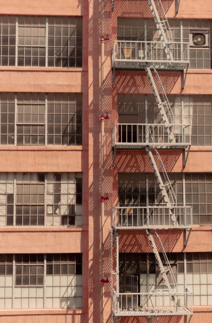 A close-up view of a multi-storey building with a brick-colored facade and large grid-style windows, some of which are open or have curtains behind them. Attached to the exterior wall is a metal fire escape staircase with several landings, supported by diagonal braces and protected by railings. The staircase runs vertically along the building, with platform sections at each floor level. Shadows from the staircase and railings cast geometric patterns onto the brick wall. The image captures part of the building's exterior environment in daylight. This scene relates to building access and safety features, relevant to house removals and moving logistics, where stair access and structural features are considerations for furniture transport and packing during a home relocation process, as managed by companies like Man With a Van Rotherhithe.