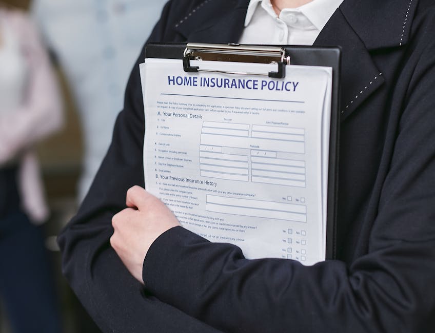 A person dressed in formal business attire, including a dark suit and white shirt, holding a clipboard with a home insurance policy form attached. The individual is gripping the clipboard with their left hand, which is partially visible, while the right hand rests against their body. The form on the clipboard features printed text, blank fields for personal details, and checkboxes, and is titled 'Home Insurance Policy.' The background appears slightly blurred, suggesting an indoor environment, possibly an office or a setting related to house removals or relocation services. This image relates to the process of home relocating and the importance of insurance and liability coverage, as referenced on the page about insurance and liability for Rotherhithe removals at manwithavanrotherhithe.co.uk, emphasizing the significance of proper documentation and protection during a move.