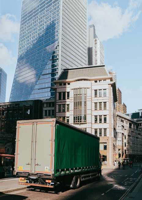 A large green and white delivery truck is parked on a city street during daytime, surrounded by tall modern glass skyscrapers and older stone buildings. The truck's rear doors are closed, and it is positioned near a sidewalk where a few pedestrians are visible in the background. The loading area shows no visible activity, with the van ready for a home relocation or furniture transport process. Bright natural light highlights the reflective surfaces of the skyscrapers and the clean, organized appearance of the moving vehicle, which is associated with professional removals services like those offered by Man With a Van Rotherhithe. The scene captures urban logistics involved in packing and moving household items in a city setting, supporting the importance of reliable removals for house relocations in Rotherhithe.