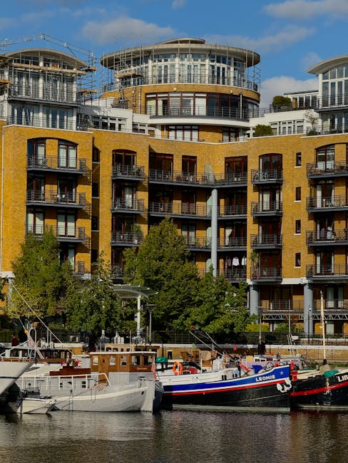 A row of boats moored along a riverbank in Rotherhithe, with a modern, multi-storey residential building featuring a yellow brick facade, numerous balconies, and large glass windows in the background. The boats are varying in size and type, some with visible equipment and outdoor furniture, while a few boats display names such as 'Leonid' and 'Liberty.' The scene is captured during daylight with clear weather, and the residential building is partially obscured by mature green trees, which provide a natural contrast to the urban setting. This image reflects a home relocation or moving environment, with the proximity of boats and a riverside property typical for moving logistics in the Rotherhithe area, supported by the services of Man With a Van Rotherhithe.