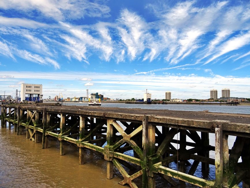 View of a wooden pier extending over a river with visible moss and algae on the supporting beams. In the background, a cityscape with several modern high-rise buildings, industrial structures, and a few smaller buildings is visible across the water. The sky above is partly cloudy with streaks of white clouds against a blue sky, and the water beneath the pier appears calm. This scene is associated with a location where house removals and furniture transport could take place, highlighting the logistics involved in moving household items near waterfront properties, as services offered by companies like Man With a Van Rotherhithe often include loading, transporting, and unloading furniture and boxes in such environments.
