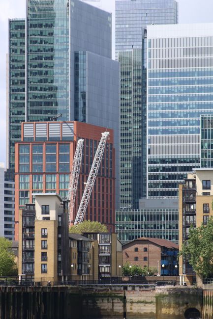 A row of boats moored along a riverbank in Rotherhithe, with a modern, multi-storey residential building featuring a yellow brick facade, numerous balconies, and large glass windows in the background. The boats are varying in size and type, some with visible equipment and outdoor furniture, while a few boats display names such as 'Leonid' and 'Liberty.' The scene is captured during daylight with clear weather, and the residential building is partially obscured by mature green trees, which provide a natural contrast to the urban setting. This image reflects a home relocation or moving environment, with the proximity of boats and a riverside property typical for moving logistics in the Rotherhithe area, supported by the services of Man With a Van Rotherhithe.