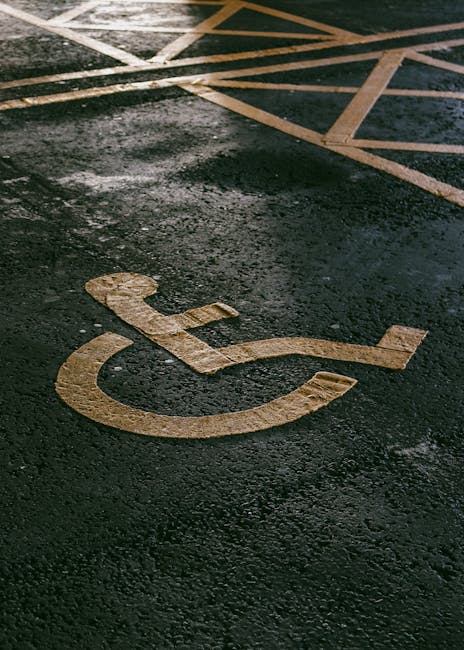 A close-up view of a black asphalt parking area featuring a yellow-painted wheelchair accessible symbol with a stylized figure using a mobility aid. In the background, there are diagonal white and yellow lines marking designated loading and parking bays, indicating a space reserved for deliveries or home relocation activities. The surface appears slightly wet, reflecting light, which enhances the visibility of the markings. This scene reflects an accessible parking zone adjacent to a property, with the surrounding environment likely intended for residential or commercial moving services. Occasionally, Man With a Van Rotherhithe provides removals and furniture transport in such areas, ensuring smooth loading and unloading within designated zones.