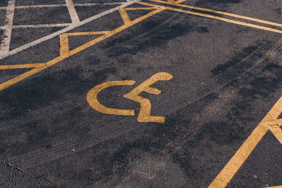 A close-up view of an accessible parking bay marked with a yellow painted wheelchair symbol on dark asphalt pavement. The parking space is set beside other marked bays, only partially visible, with yellow lines outlining their boundaries. The surface appears clean but shows some texture and tire marks from vehicle movement. The area is outdoors, underground or open-air, with natural lighting. There are no vehicles or objects within the parking bay. This image highlights accessible parking provisions relevant to home relocation and furniture transport processes, as provided by Man With a Van Rotherhithe, situated in a typical urban setting near residential or commercial premises, supporting efficient loading and unloading during house removals or moving services.