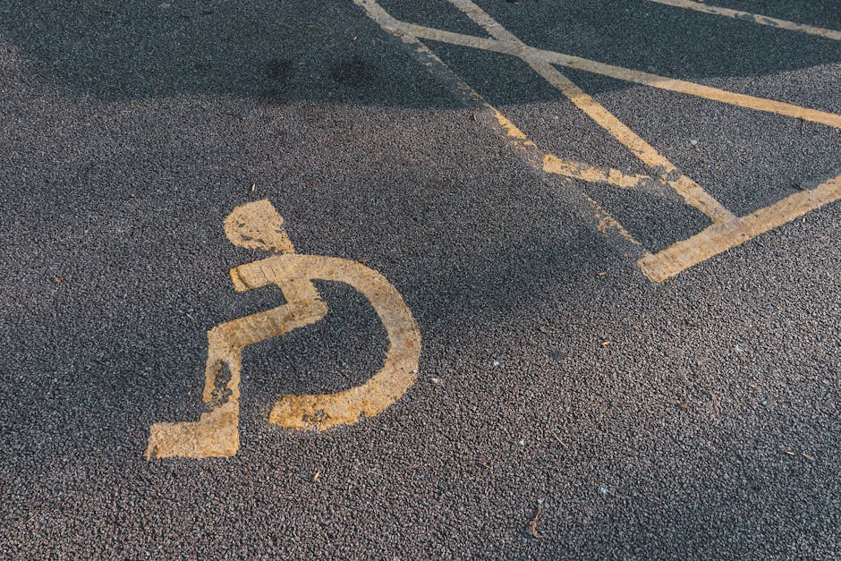 A close-up view of a black asphalt parking area featuring a yellow-painted wheelchair accessible symbol with a stylized figure using a mobility aid. In the background, there are diagonal white and yellow lines marking designated loading and parking bays, indicating a space reserved for deliveries or home relocation activities. The surface appears slightly wet, reflecting light, which enhances the visibility of the markings. This scene reflects an accessible parking zone adjacent to a property, with the surrounding environment likely intended for residential or commercial moving services. Occasionally, Man With a Van Rotherhithe provides removals and furniture transport in such areas, ensuring smooth loading and unloading within designated zones.