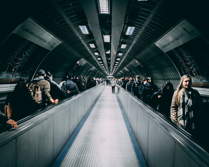 A busy underground pedestrian tunnel with a curved, metallic ceiling featuring rectangular lighting panels. The tunnel's sides are lined with advertising banners. On each side of a moving walkway, numerous people are standing and walking, some carrying bags or backpacks. The floor consists of a textured, non-slip surface, with the moving walkway in the centre. The tunnel appears well-lit, with a mix of natural and artificial light, providing a clear view of the individuals and the surrounding environment. This scene depicts a typical urban passageway, commonly used by commuters and travellers, illustrating the kind of environment where home relocation or furniture transport could involve navigating through tight or tricky access points, similar to the challenges addressed by removal services like Man With a Van Rotherhithe.