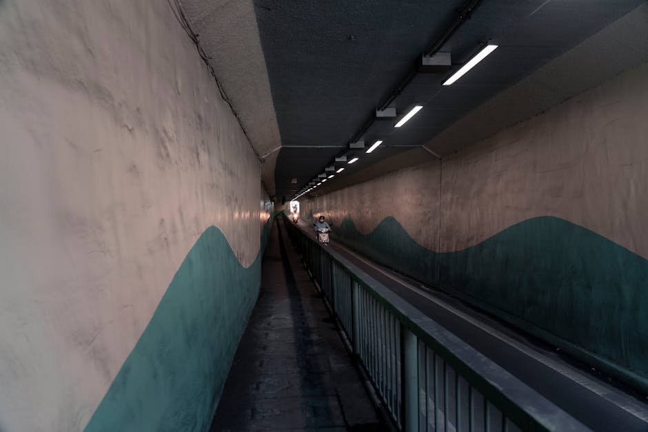 A construction worker wearing a red safety helmet is seen smiling through an open walkway within a large underground tunnel boring machine, which has a circular cutting head with metal teeth and detailed mechanical components. The tunnel walls are lined with metal reinforcement bars and support structures, and the surrounding environment is dimly lit with visible cables and scaffolding. The worker appears to be inside a confined space, surrounded by concrete and steel, indicating a segment of the home relocation process involving heavy machinery and precise positioning of equipment. This scene illustrates the complex logistics and machinery involved in underground construction or tunneling projects, relevant to the scope of house removals and transport services, especially when navigating tricky access jobs such as tunnel or excavation work. Man With a Van Rotherhithe offers professional removals and logistical support for projects that require careful planning and equipment handling during home or property relocations.