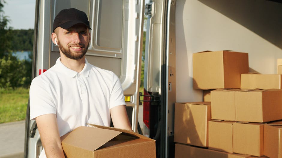 A white moving van parked on a city street with a man with long hair, wearing a headband and a blue blazer, leaning out of the driver's side window. The van displays the company name 'Moving Company'