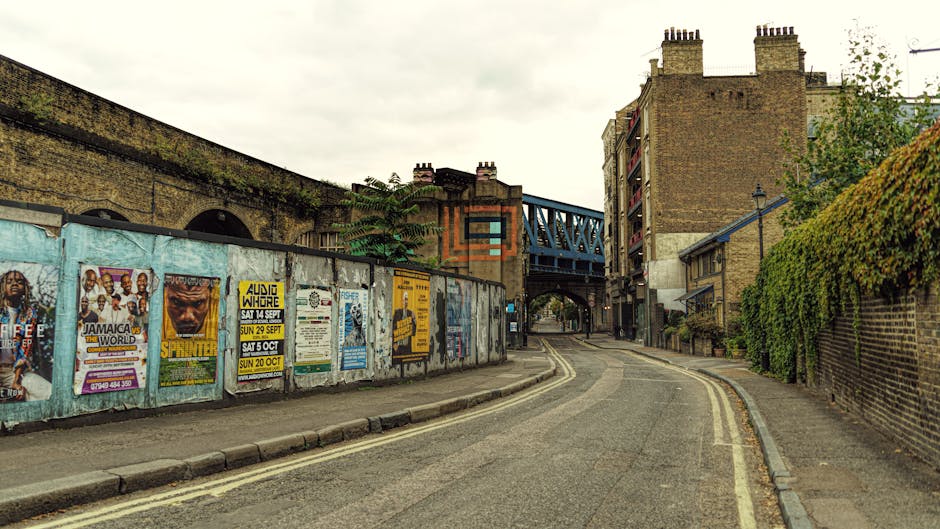 A narrow residential street in Bermondsey Wall and Greenland Dock area during daytime, with an overcast sky. The scene shows a gently curved tarmac road with double yellow lines on the left side, leading under a blue metal bridge structure with visible supports. On the left, there is a long brick wall with weathered posters and advertisements featuring event promotions, partially covered with graffiti and peeling paint. On the right, a brick building with balconies and chimney stacks is adjacent to a green hedge running along the sidewalk, which appears to be part of a property boundary. The street is devoid of traffic or pedestrians, and the lighting is natural and diffuse, suggesting cloudy weather. This setting captures a typical urban environment where residential property relocation or moving services might be carried out, with trucks or vans potentially parked nearby for furniture transport or home relocation activities as provided by Man With a Van Rotherhithe.