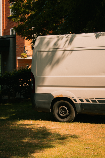 A beige moving van parked on a grassy area adjacent to a residential building, partially shaded by overhanging tree branches. The van’s rear end and side are visible, with black trim and a few rust marks near the wheel arch. In the background, there are red-brick apartment buildings with white window frames, and a small shrubbery near the vehicle. The scene captures the loading or unloading process typical of house removals, suggesting furniture or boxes may be transported. Man With a Van Rotherhithe is a professional removal service that facilitates home relocation and furniture transport, often involving vehicles like this in their packing and moving operations.