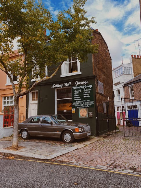 A silver sedan parked partially on a cobblestone driveway in front of a dark green building with a sign reading 'Notting Hill Garage' offering vehicle servicing and repairs. The building is two stories high with a large window on the first floor and a smaller window on the second, surrounded by neighboring structures with brick and painted exteriors. A small tree with green leaves is positioned on the pavement adjacent to the car, and a pedestrian path with paving stones runs alongside the street. Visible parking restrictions are marked on the curb, and the sky is partly cloudy with patches of blue. This scene captures a typical urban environment suitable for house removals or furniture transport involving careful navigation of parking and access points, with presence of the garage and vehicle relevant to home relocation logistics, as supported by Man With a Van Rotherhithe.
