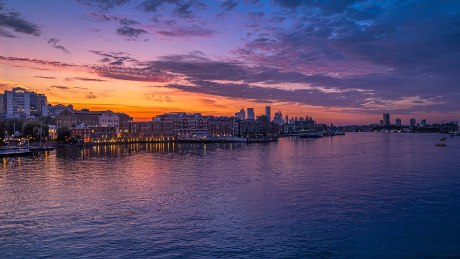 A scenic view of the River Thames at sunset, with a skyline of modern and historic buildings along the waterfront. The sky is painted with shades of orange, pink, and purple, reflecting softly on the water's surface. In the foreground, the river's calm waters gently ripple, with a few small boats visible in the distance. The cityscape includes a mix of mid-rise structures and taller towers, suggesting urban development near the water. This image captures the peaceful yet vibrant atmosphere of the Surrey Quays to Deptford area, highlighting the importance of efficient home relocation and furniture transport logistics, as demonstrated by professional removals services such as those provided by Man With a Van Rotherhithe.