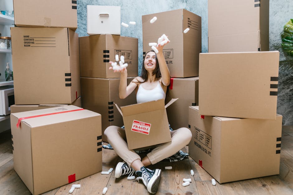 A young woman sits cross-legged on a wooden floor inside a room, surrounded by numerous large cardboard boxes, some sealed with red tape and others labeled with 'fragile'. She is wearing a white tank