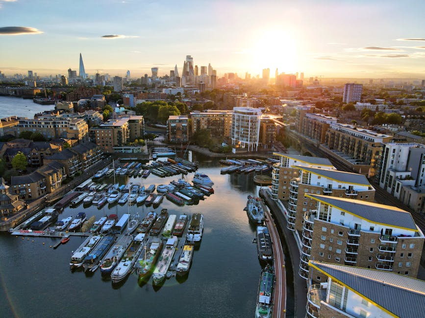 Aerial view of Rotherhithe in London during sunset, showing a marina with numerous boats docked along the pier and surrounding modern residential buildings. The scene includes a mix of wooden and concrete structures, with some boats partially covered in protective plastic and others uncovered. Visible on the scene are boat slips, wharves, and pathways along the water's edge. In the background, the iconic London skyline features prominent skyscrapers such as The Shard and other high-rise buildings, with the sunlight casting a warm glow over the urban landscape. This image captures the essence of home relocation and furniture transport in Rotherhithe, highlighting the busy environment where residents and professional services like [COMPANY_NAME] may facilitate removals, packing, and loading processes, especially in preparation for a same-day move or transport to new properties.