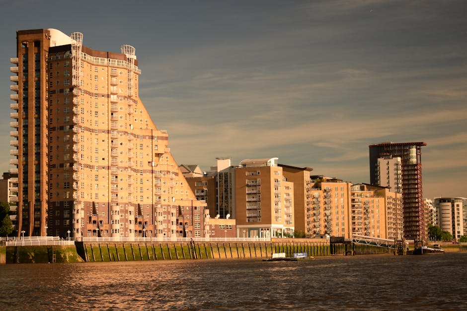 Photograph of a cityscape along a riverbank during daylight, featuring several multi-storey residential buildings with varied architectural styles and heights. The tallest building on the left has a distinctive curved design with numerous small, evenly spaced windows and balconies, constructed with brick and concrete materials. Adjacent are mid-rise apartment blocks with beige and cream facades, each with multiple balconies, some with glass railings. To the right, a partially constructed high-rise with exposed red steel framework and scaffolding is visible, indicating ongoing development. The buildings are situated on a grassy embankment with a low retaining wall and wooden fencing, while boats are moored along the river in the foreground. The sky is partly cloudy with soft, warm lighting, capturing the urban environment that could correspond to a house removal or moving context in Rotherhithe, supported by the nearby waterways and residential skyline.